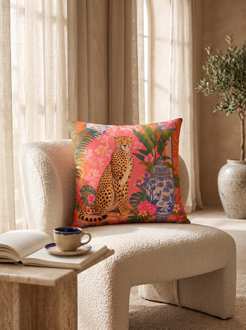 A square decorative throw pillow with a vibrant tropical print featuring a cheetah, flowers, and a blue porcelain pot, styled on a cream boucle armchair in a sunlit room.