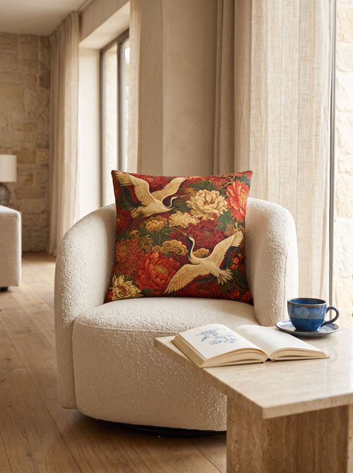 A decorative throw pillow with a red and gold floral crane pattern, styled on a cream boucle armchair in a warm, sunlit living room.