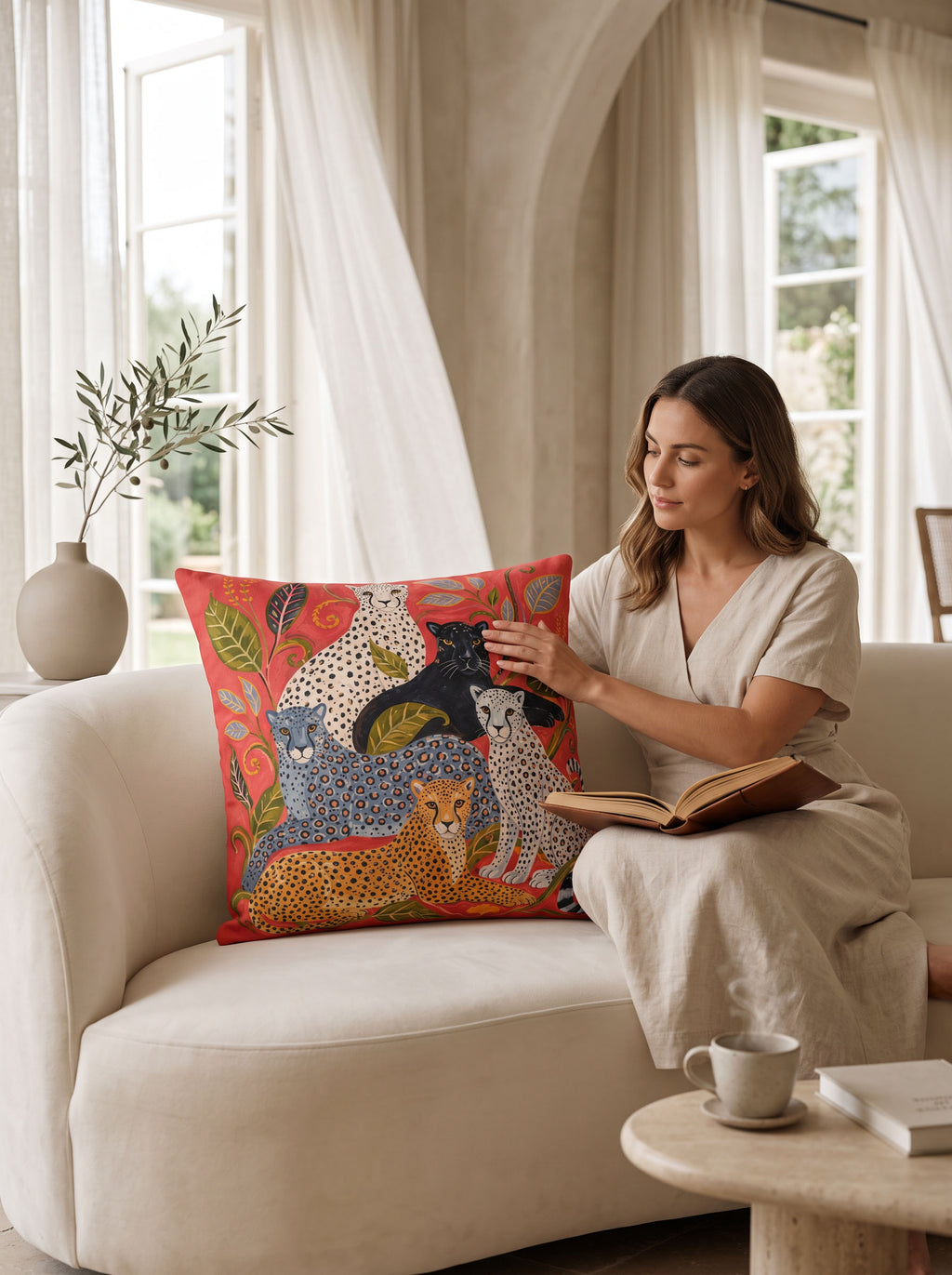 A woman sits on a cream sofa in a bright living room, holding a square decorative throw pillow with a colorful animal print. The pillow features a red-orange background with illustrated leopards, cheetahs, and other cats in blue, black, beige, and green tones.