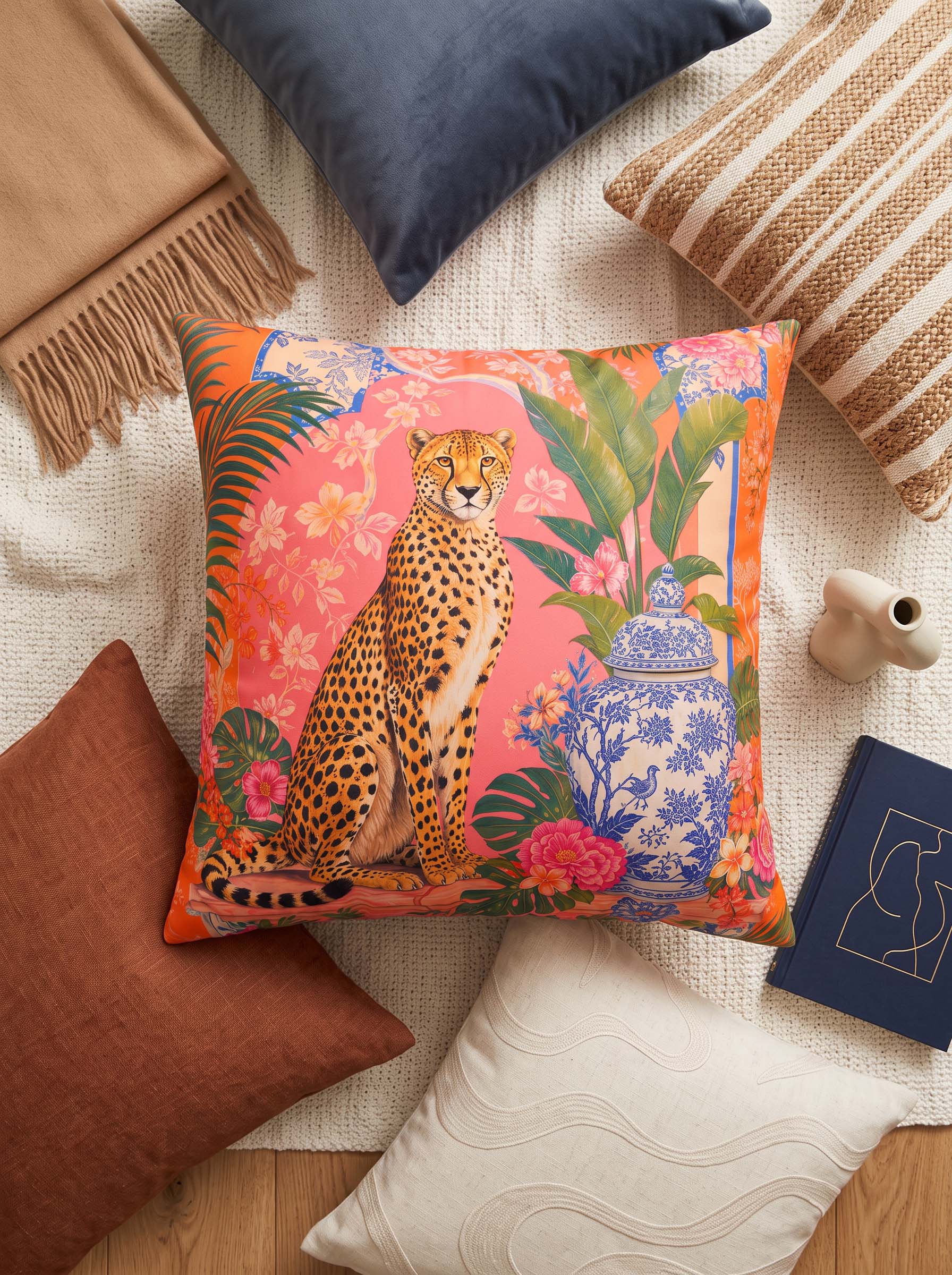 A square decorative throw pillow featuring a colorful print of a seated cheetah with tropical foliage, pink flowers, and a blue-and-white porcelain vase on a coral background. The pillow is styled in a cozy flat-lay arrangement with other pillows and home decor props on a cream textured blanket.