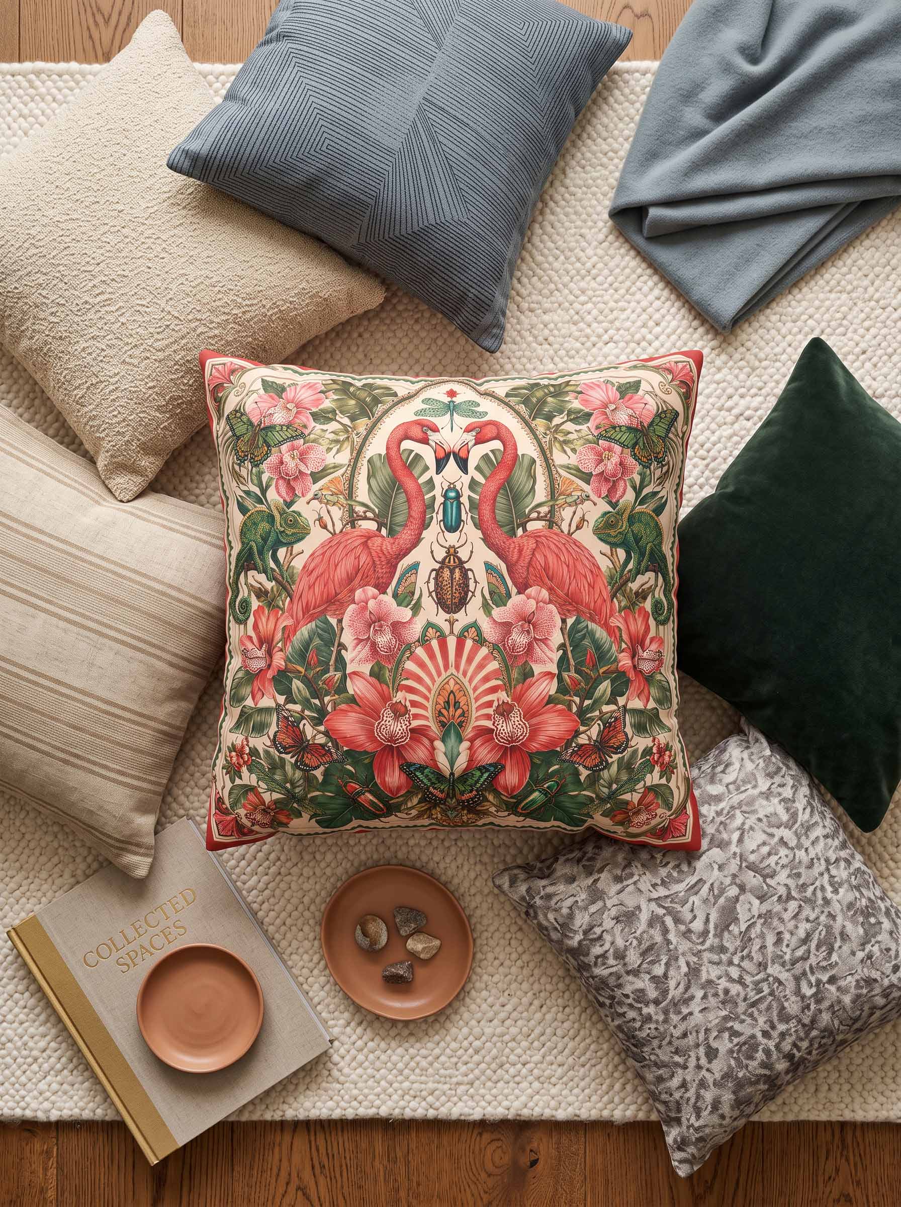 A decorative square throw pillow with a detailed Art Nouveau-style flamingo and floral pattern in pink, red, green, and cream, styled in a cozy flat-lay arrangement with other pillows, a blanket, and books on a woven rug.