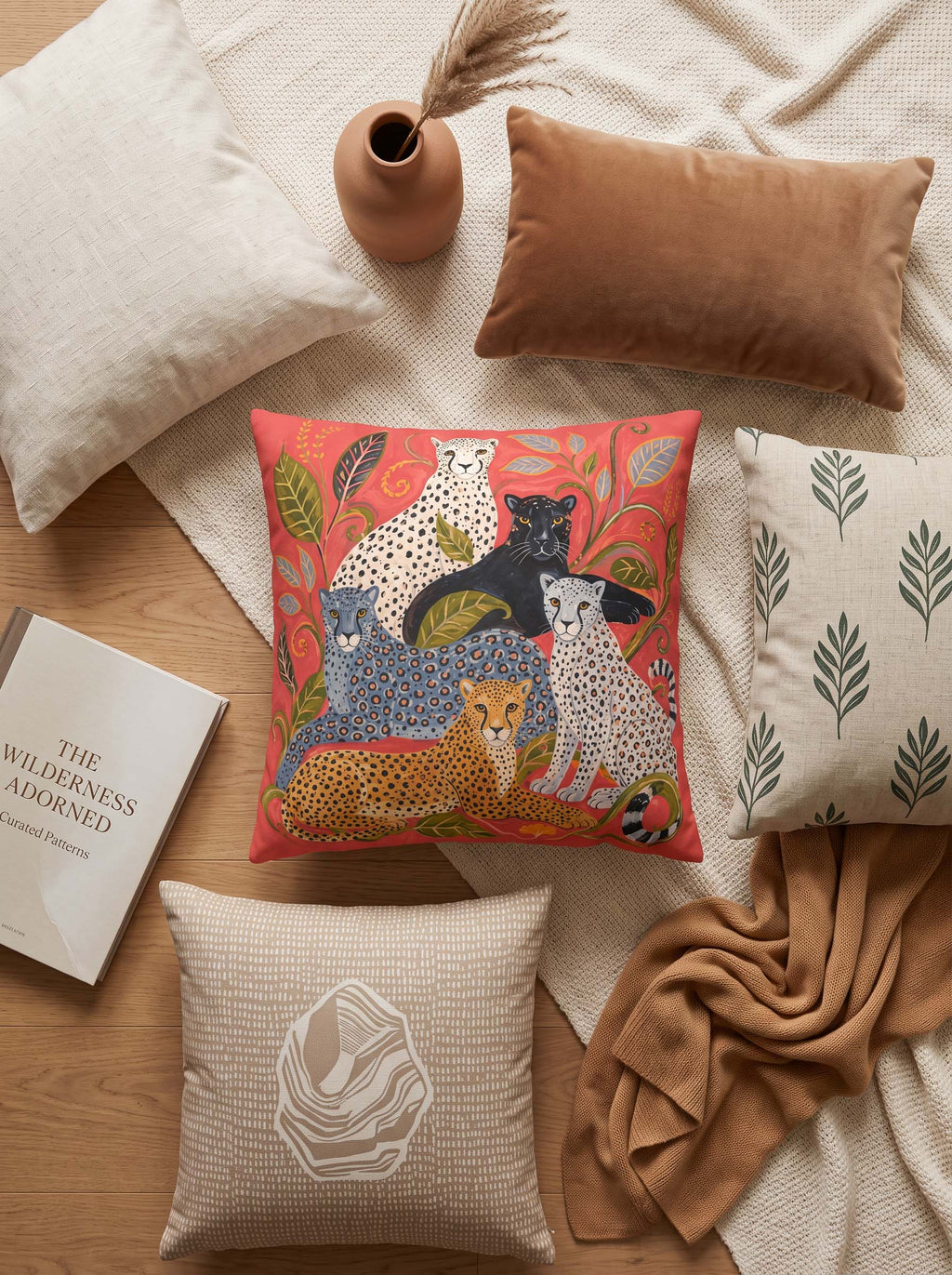 A styled flat-lay of decorative throw pillows on a wood floor with neutral textiles and props. The central product is a square pillow featuring a colorful safari-style illustration of spotted cats and a leopard on a coral-red background.